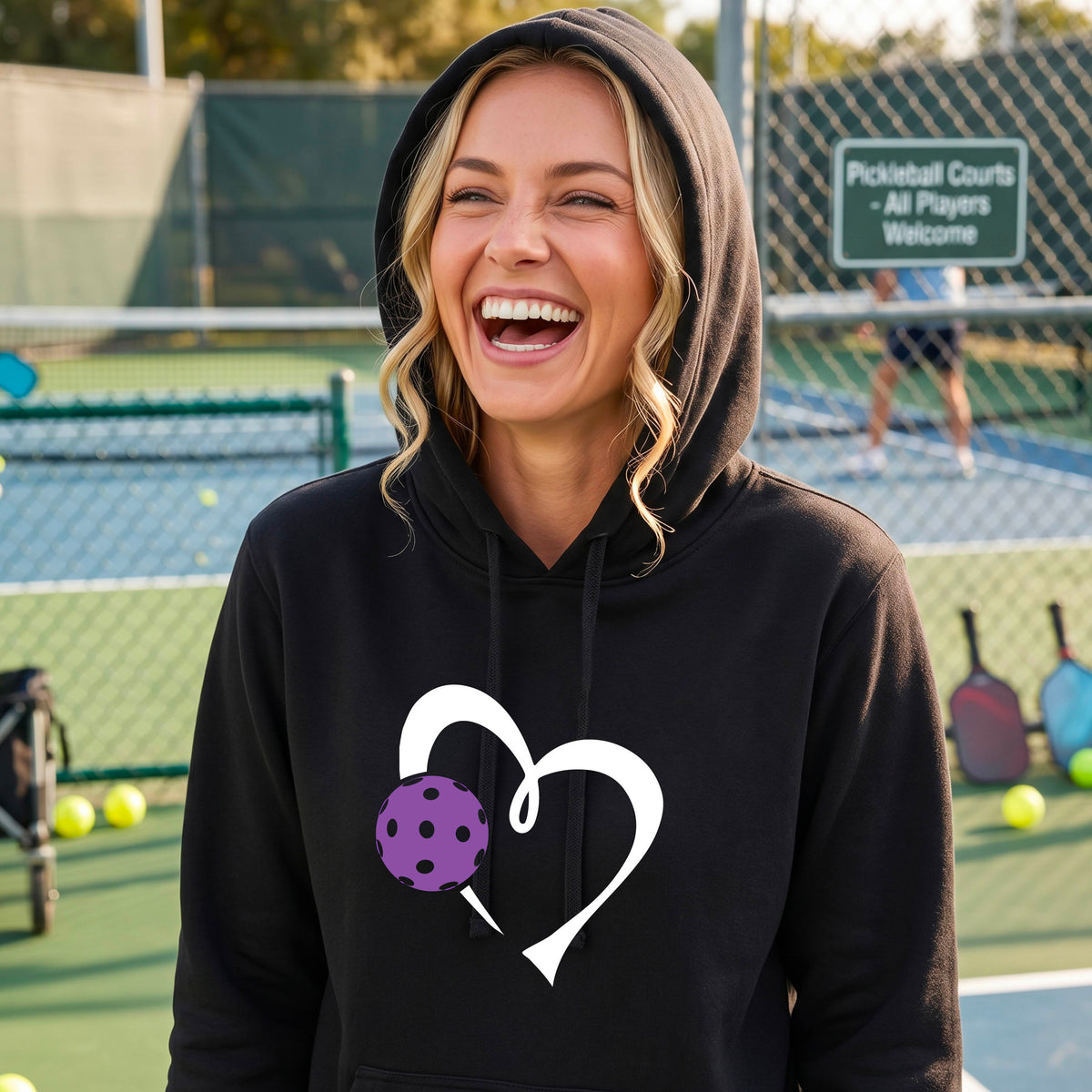 A woman smiling on an outdoor pickleball court wearing a black hoodie with a white heart outline and a purple pickleball graphic on the front. 