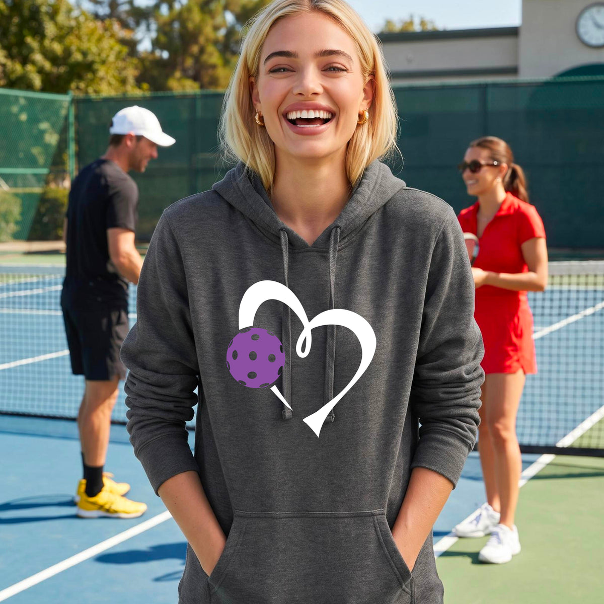A woman smiling on an outdoor pickleball court wearing a dark gray hoodie with a white heart outline and a purple pickleball graphic on the front. 