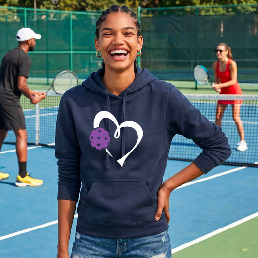 A woman smiling on an outdoor pickleball court wearing a navy blue hoodie with a white heart outline and a purple pickleball graphic on the front. 