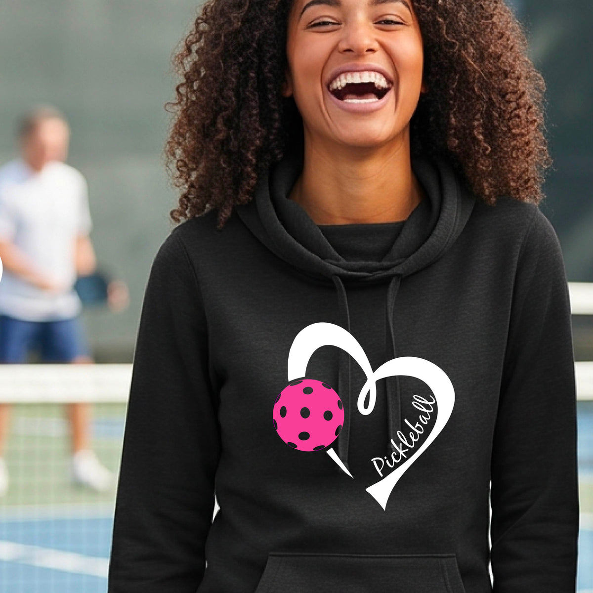 A woman wearing a black cowl-neck hoodie with a white heart outline, a pink pickleball graphic, and the word 'Pickleball' printed on the front, photographed on an outdoor tennis court.