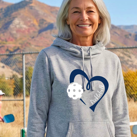 A woman wearing a light gray cowl-neck hoodie with a navy blue heart outline, a white pickleball graphic, and the word 'Pickleball' printed on the front.