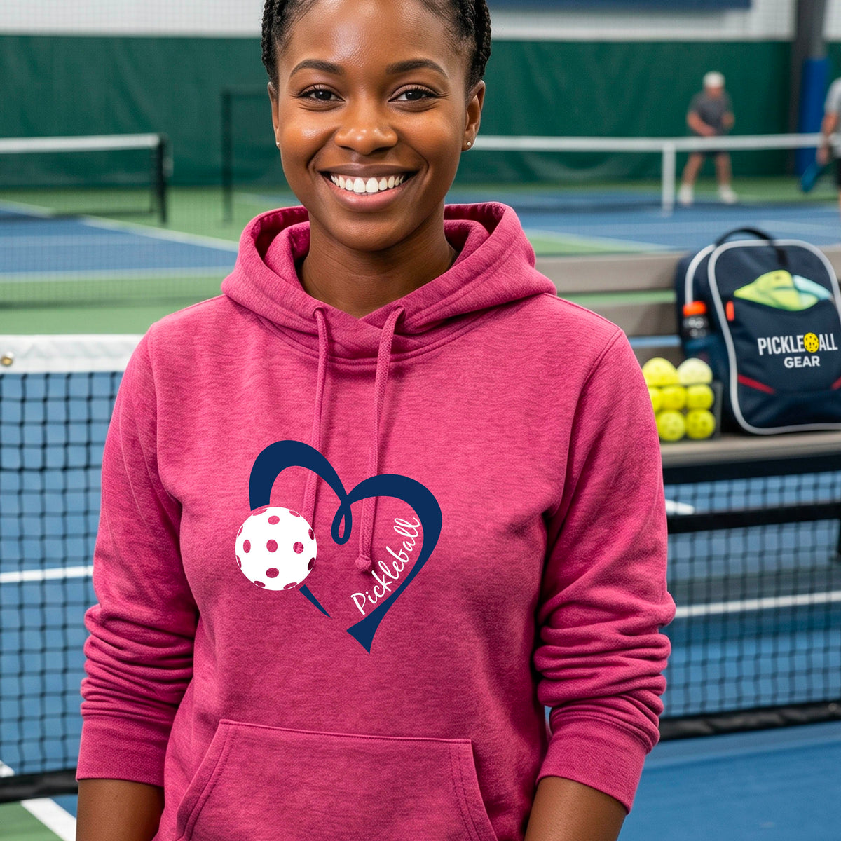 A woman wearing a pink cowl-neck hoodie with a navy blue heart outline, a white pickleball graphic, and the word 'Pickleball' printed on the front.