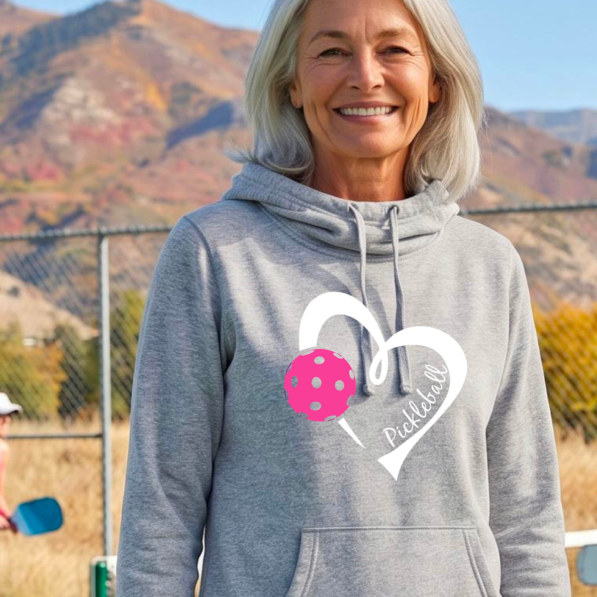 A woman wearing a light gray cowl-neck hoodie with a white heart outline, a pink pickleball graphic, and the word 'Pickleball' printed on the front, photographed on an outdoor tennis court.
