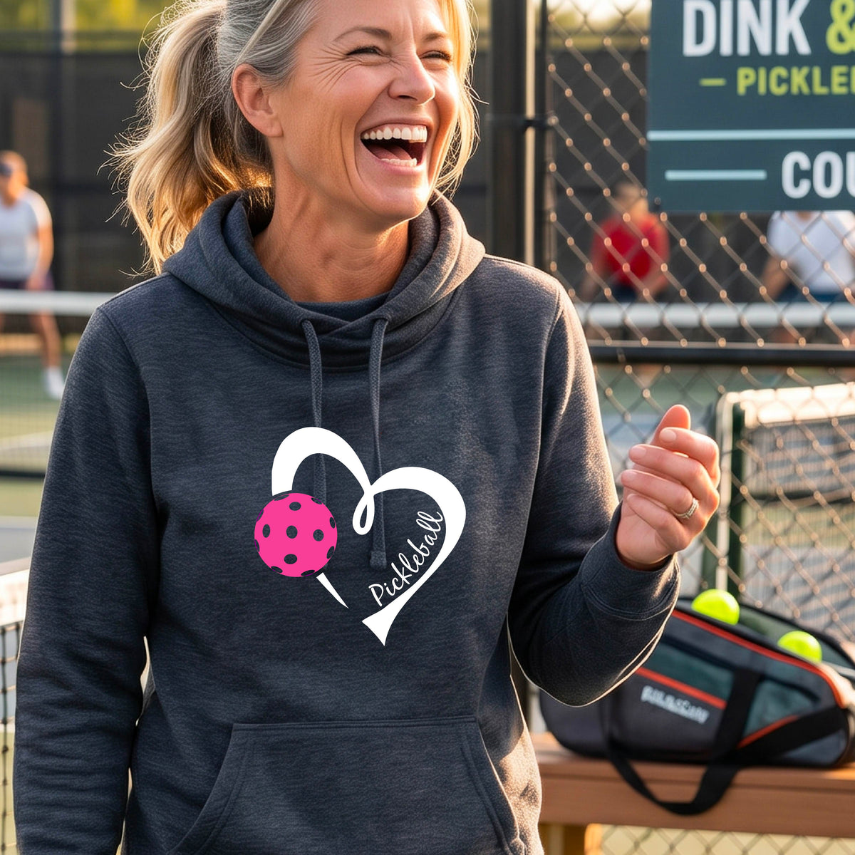A woman wearing a navy blue cowl-neck hoodie with a white heart outline, a pink pickleball graphic, and the word 'Pickleball' printed on the front, photographed on an outdoor tennis court.