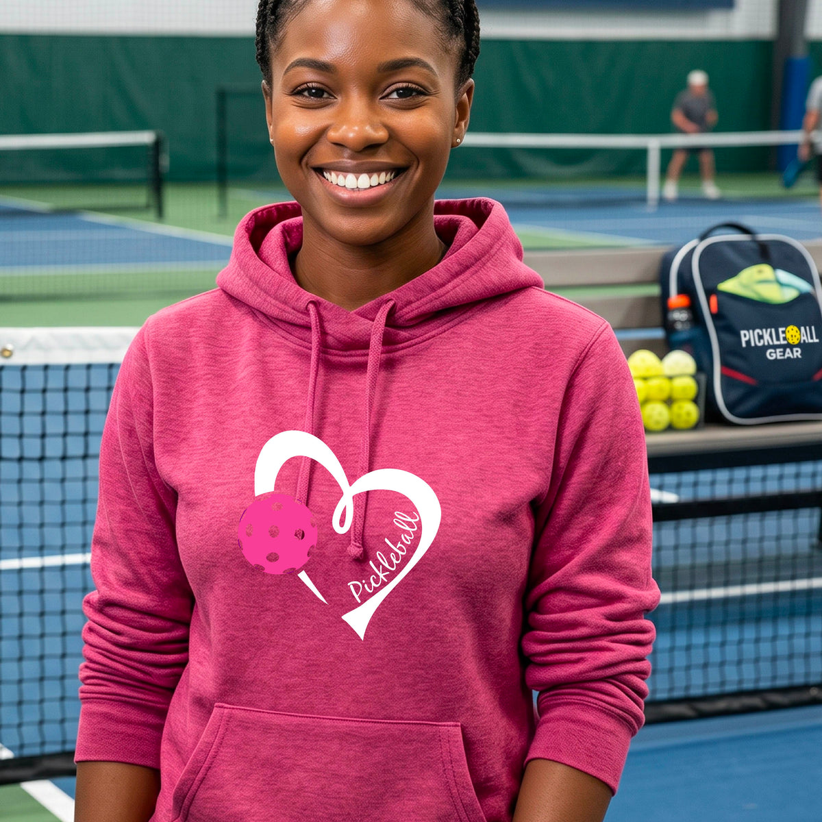 A woman wearing a pink cowl-neck hoodie with a white heart outline, a pink pickleball graphic, and the word 'Pickleball' printed on the front, photographed on an outdoor tennis court.