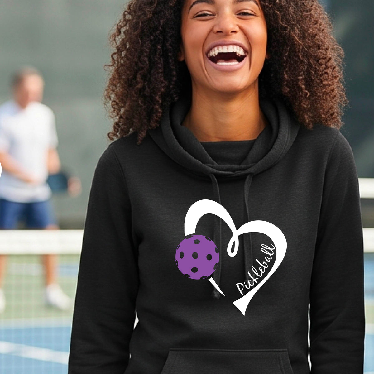 A smiling woman stands outdoors wearing a black pullover hoodie with a white heart outline and a purple pickleball graphic on the front.
