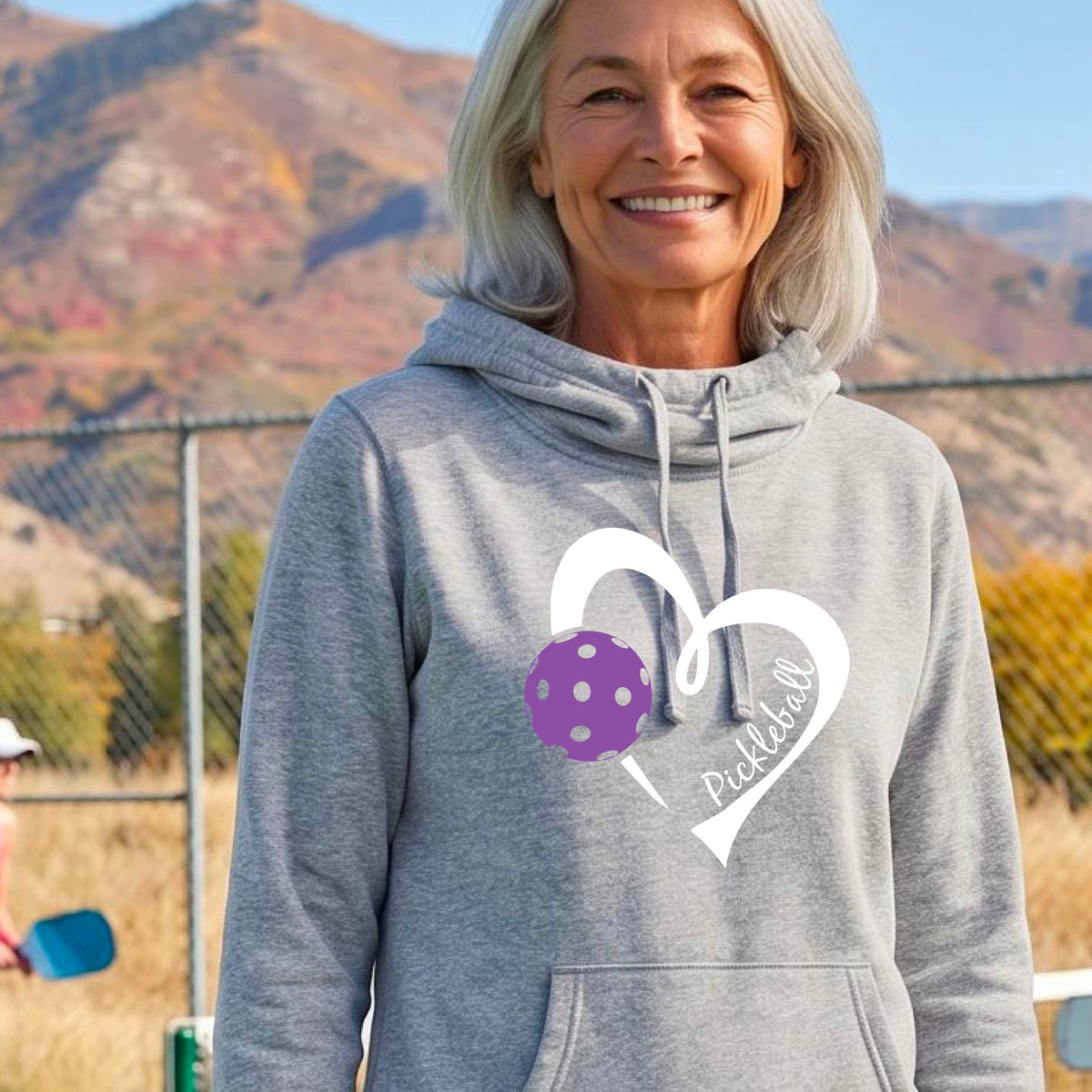A smiling woman stands outdoors wearing a heather gray pullover hoodie with a white heart outline and a purple pickleball graphic on the front.