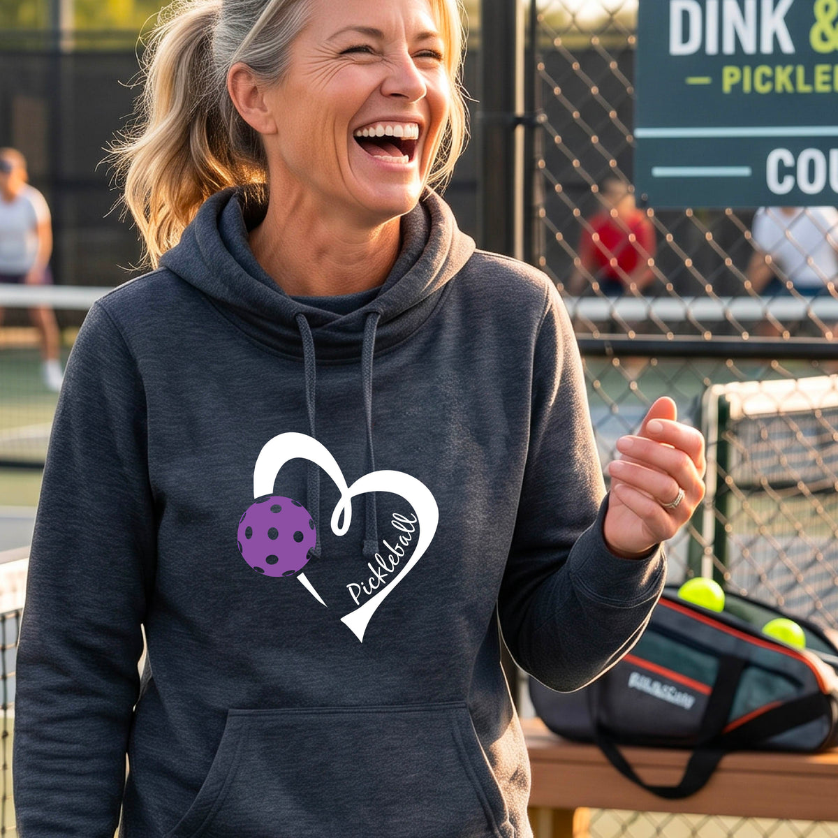 A smiling woman stands outdoors wearing a navy blue pullover hoodie with a white heart outline and a purple pickleball graphic on the front.