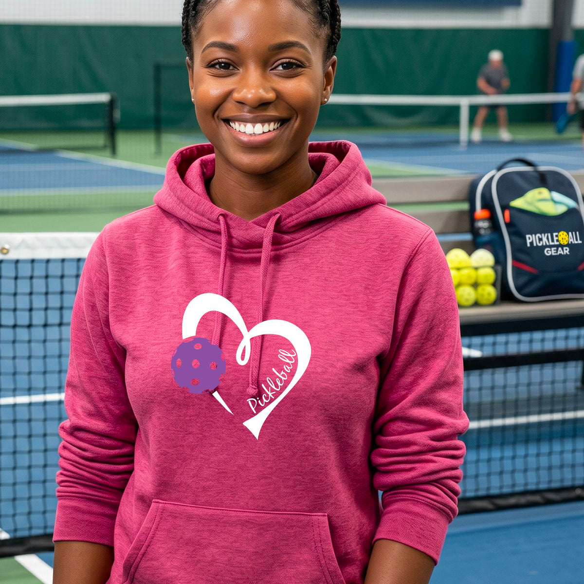 A smiling woman stands outdoors wearing a pink pullover hoodie with a white heart outline and a purple pickleball graphic on the front.