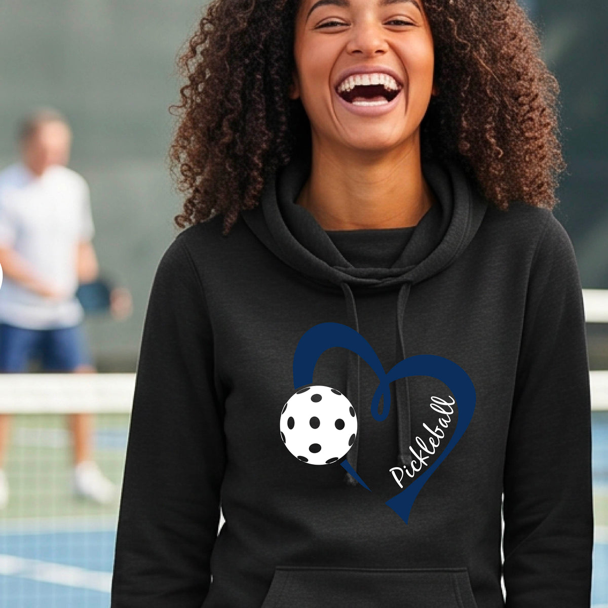 A woman wearing a black cowl-neck hoodie with a navy blue heart outline, a white pickleball graphic, and the word 'Pickleball' printed on the front.