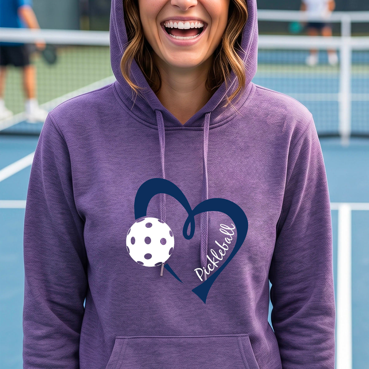 A woman wearing a purple pullover hoodie with a navy blue heart outline, a white pickleball, and the word 'Pickleball' printed on the front.