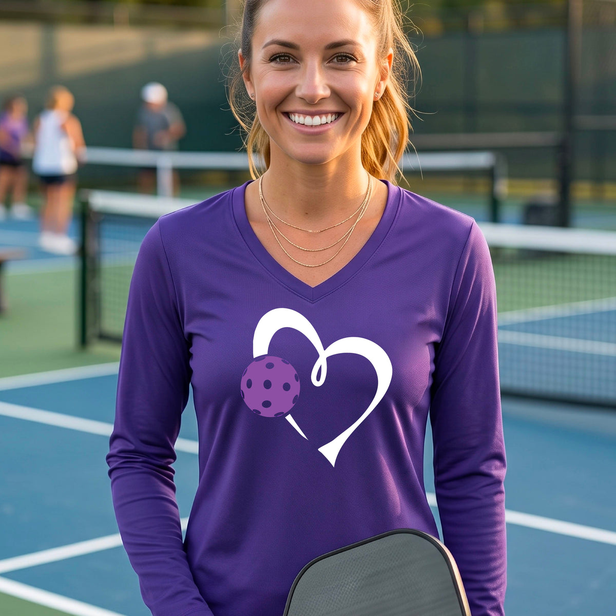 A smiling older woman on a pickleball court wearing a purple long-sleeve V-neck shirt with a white heart outline and a purple pickleball graphic. 