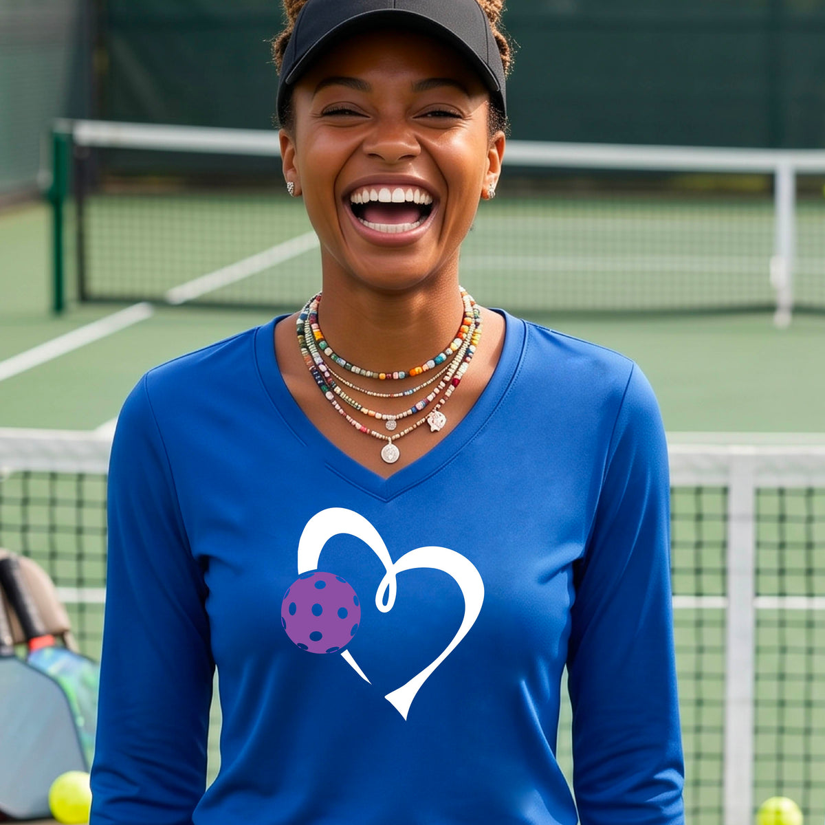 A smiling older woman on a pickleball court wearing a bright royal blue long-sleeve V-neck shirt with a white heart outline and a purple pickleball graphic. 
