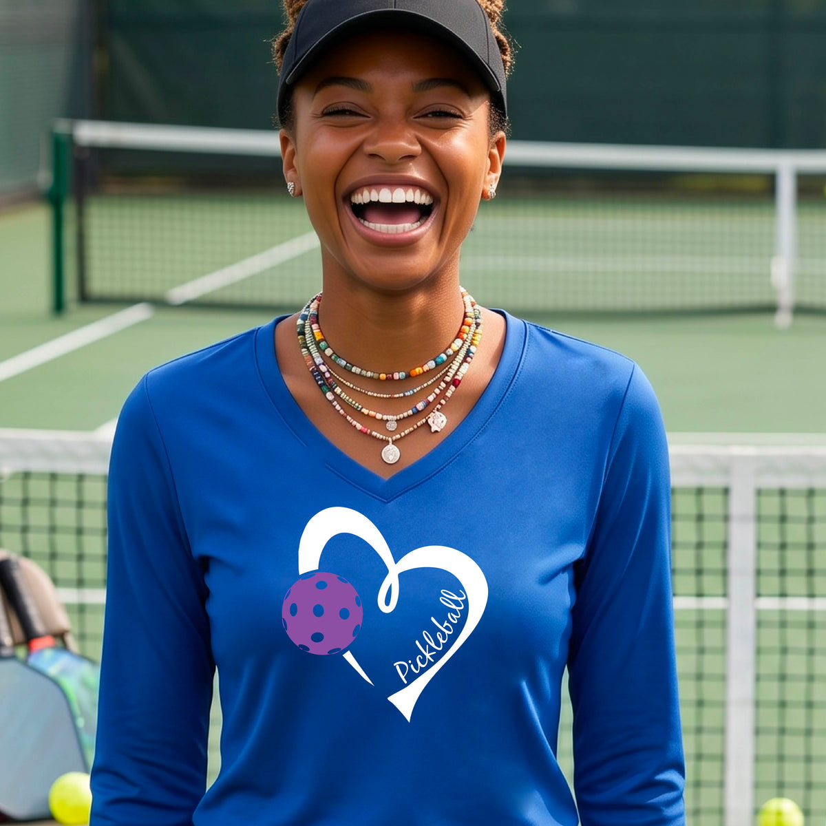 A smiling woman stands on a pickleball court wearing royal blue long-sleeve V-neck shirt with a white heart outline, a purple pickleball graphic, and the word 'Pickleball' printed on the front. 