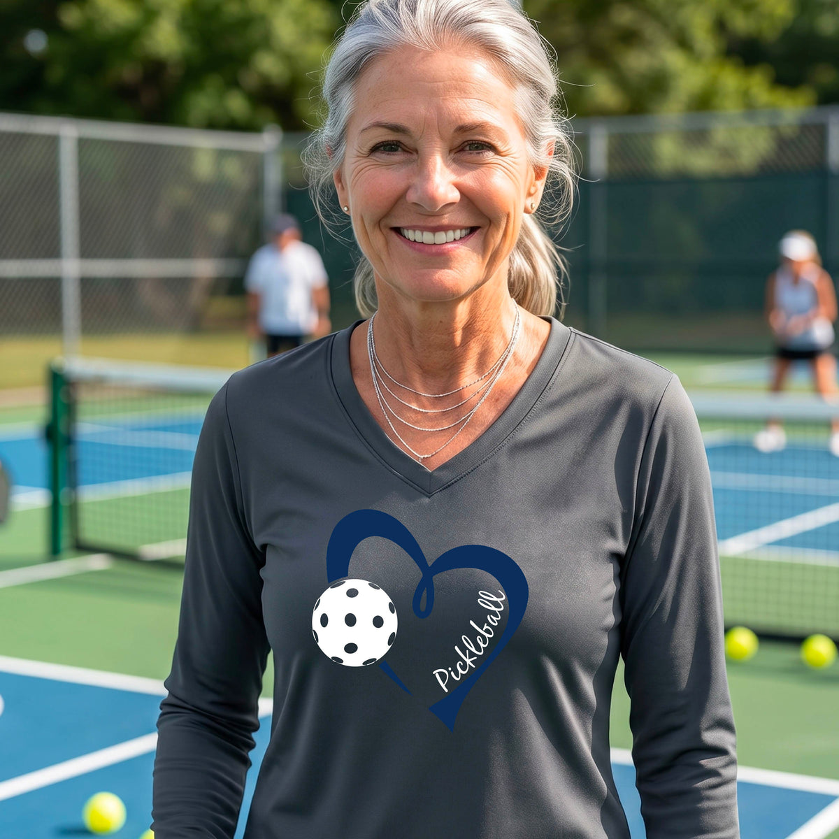 A woman on a pickleball court wearing a dark gray long-sleeve V-neck shirt with a dark blue heart graphic, a white pickleball ball, and the word 'Pickleball' on the front.