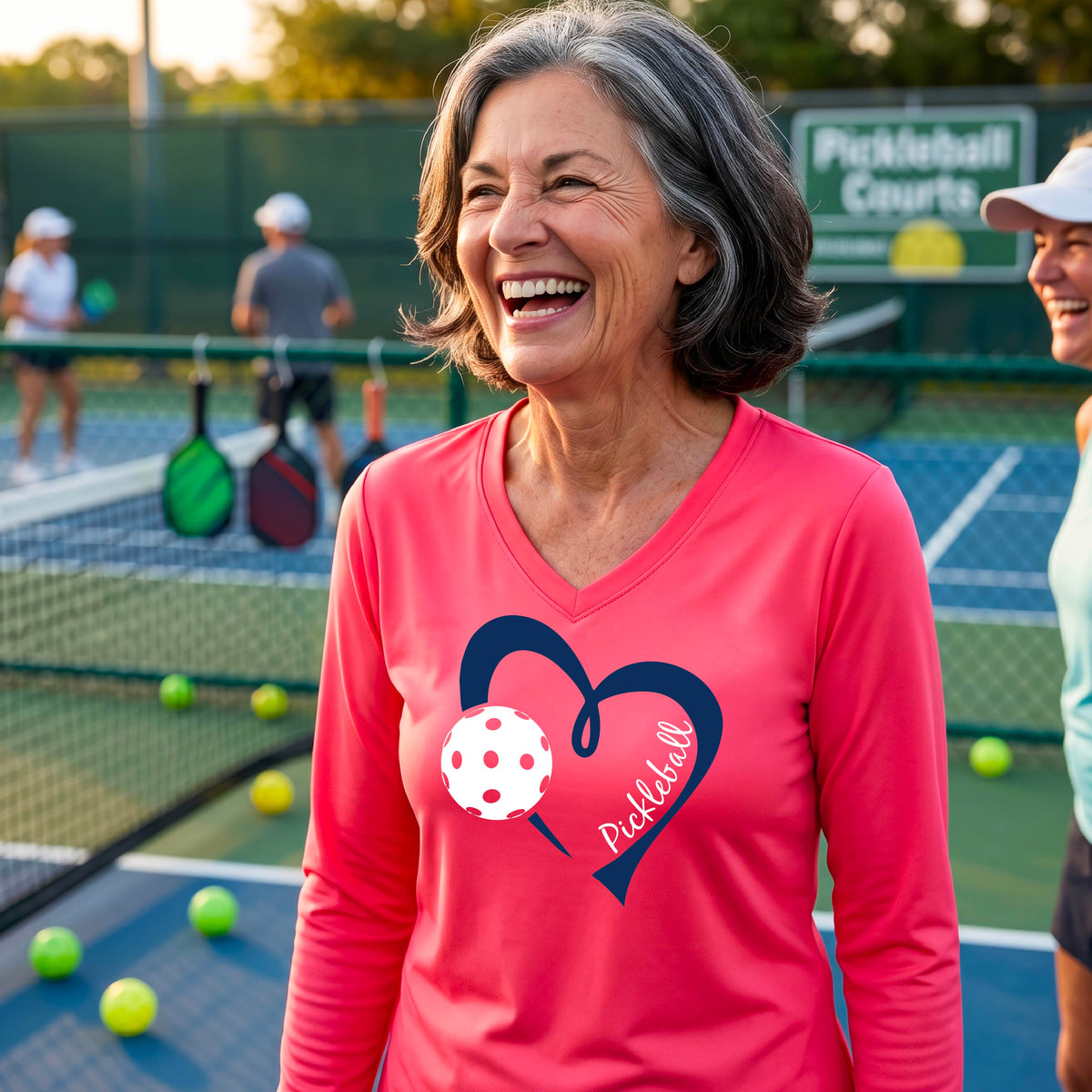 A woman on a pickleball court wearing a coral pink long-sleeve V-neck shirt with a dark blue heart graphic, a white pickleball ball, and the word 'Pickleball' on the front.