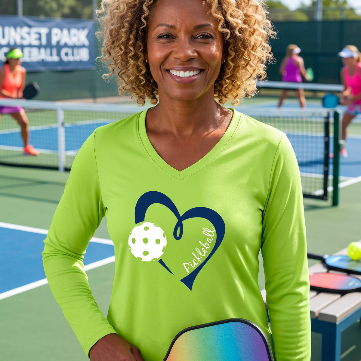 A woman on a pickleball court wearing a lime green long-sleeve V-neck shirt with a dark blue heart graphic, a white pickleball ball, and the word 'Pickleball' on the front.