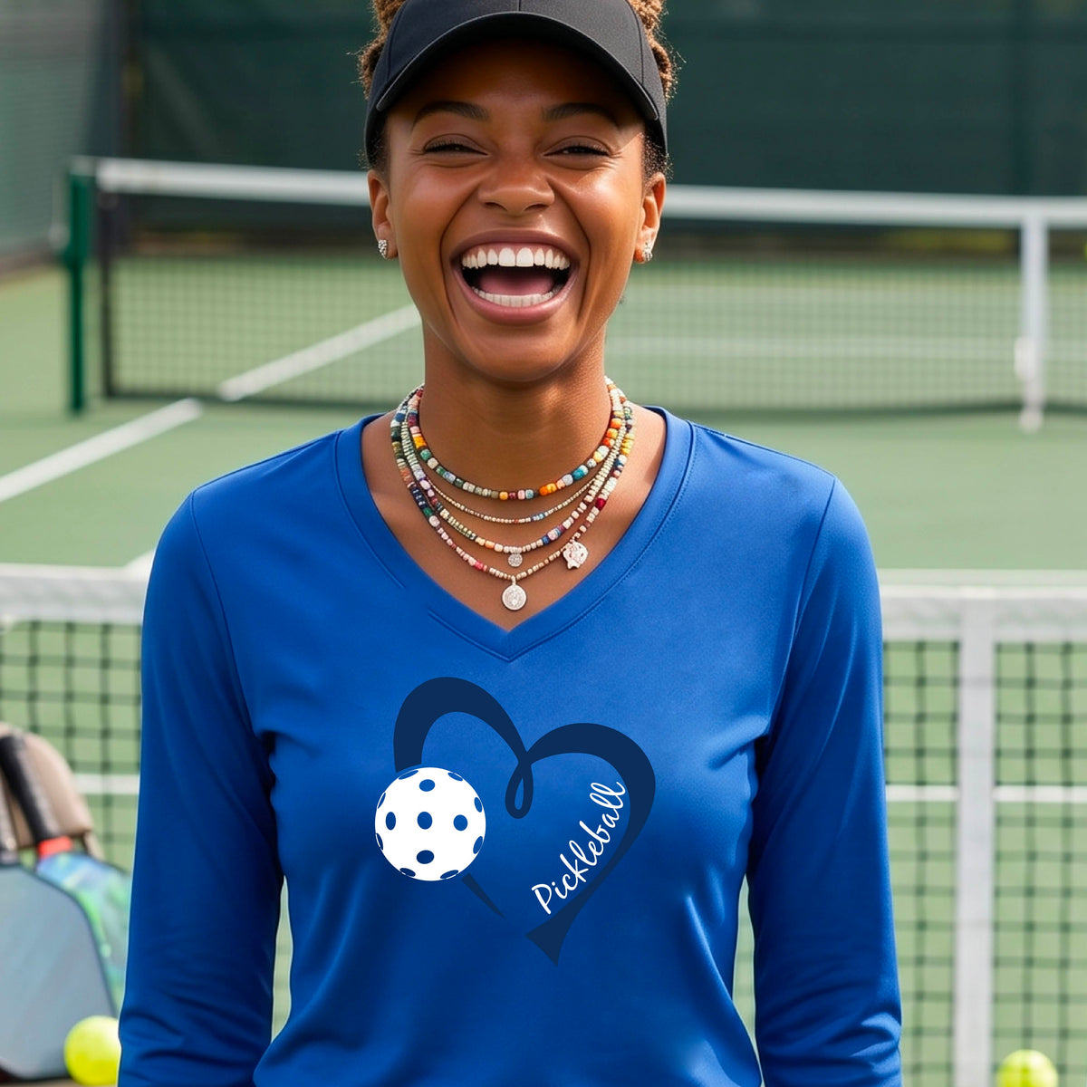 A woman on a pickleball court wearing a bright royal blue long-sleeve V-neck shirt with a dark blue heart graphic, a white pickleball ball, and the word 'Pickleball' on the front.