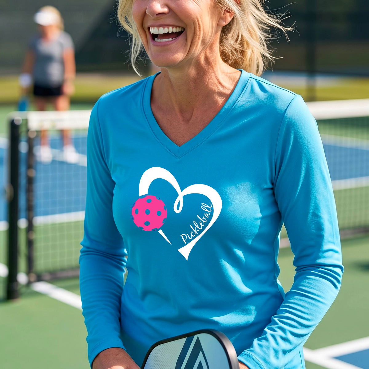 A woman smiling on a pickleball court wearing a bright light blue long-sleeve V-neck athletic shirt with a white heart outline, pink pickleball graphic, and the word 'Pickleball' printed on the front. 