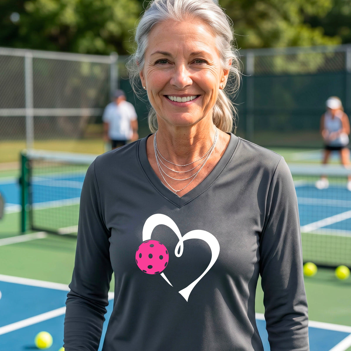 A smiling woman on a pickleball court wearing a dark gray long-sleeve V-neck shirt with a white heart outline and a pink pickleball graphic on the front.