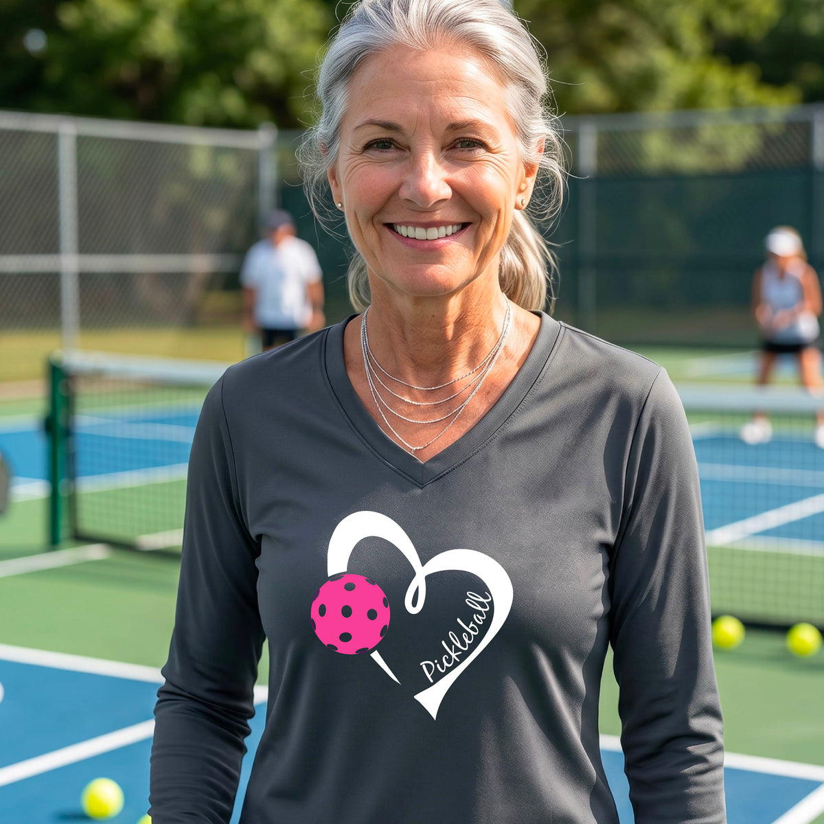 A woman smiling on a pickleball court wearing a dark gray long-sleeve V-neck athletic shirt with a white heart outline, pink pickleball graphic, and the word 'Pickleball' printed on the front. 