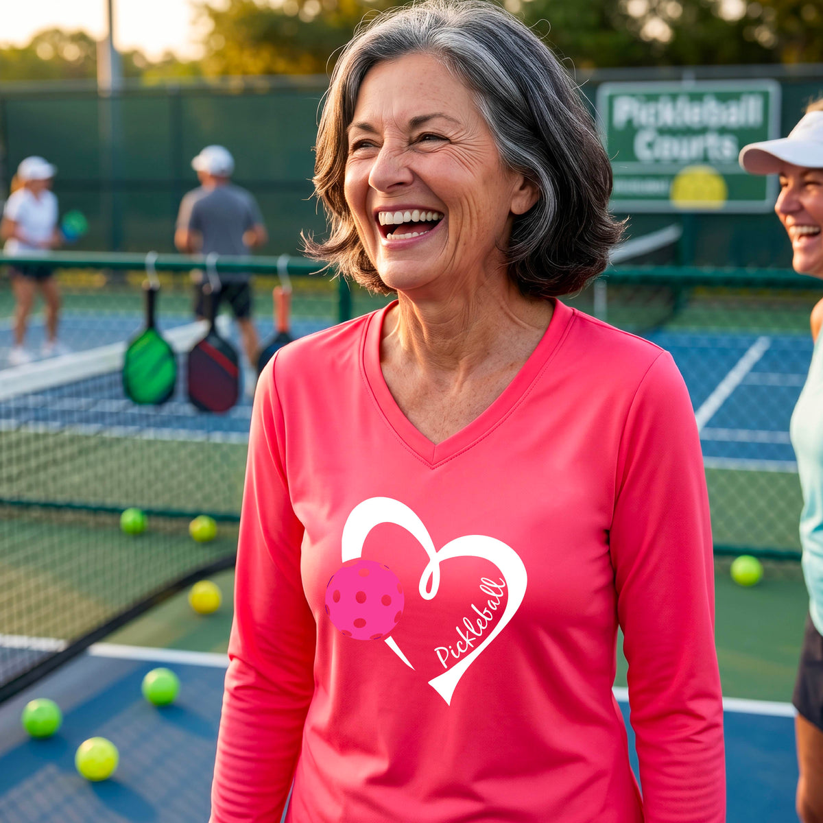 A woman smiling on a pickleball court wearing a bright coral pink long-sleeve V-neck athletic shirt with a white heart outline, pink pickleball graphic, and the word 'Pickleball' printed on the front. 