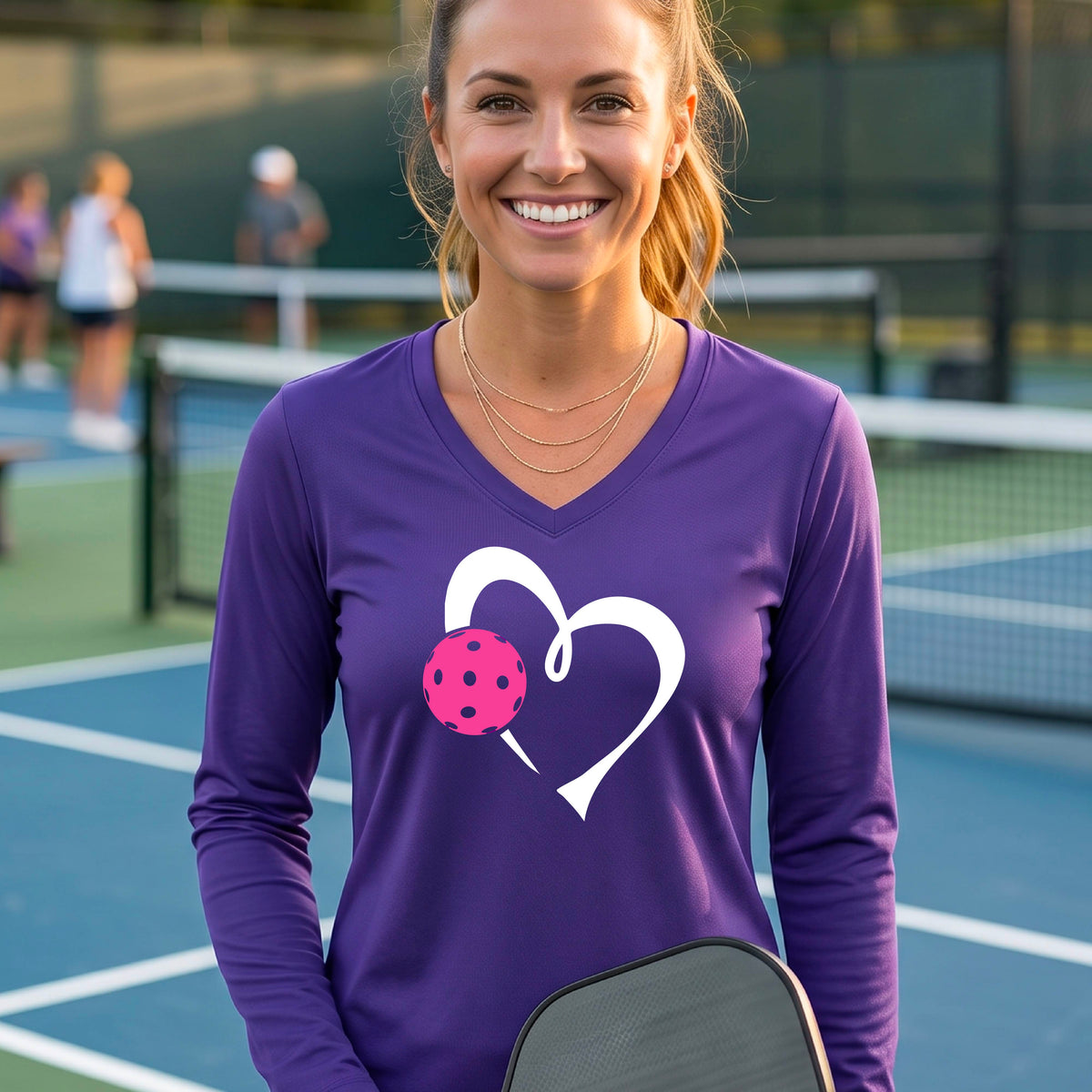 A smiling woman on a pickleball court wearing a purple long-sleeve V-neck shirt with a white heart outline and a pink pickleball graphic on the front.