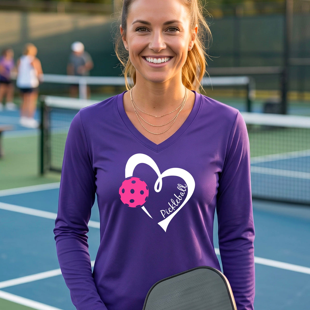 A woman smiling on a pickleball court wearing a purple long-sleeve V-neck athletic shirt with a white heart outline, pink pickleball graphic, and the word 'Pickleball' printed on the front. 