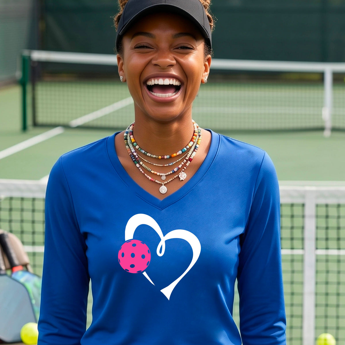 A smiling woman on a pickleball court wearing a royal blue long-sleeve V-neck shirt with a white heart outline and a pink pickleball graphic on the front.