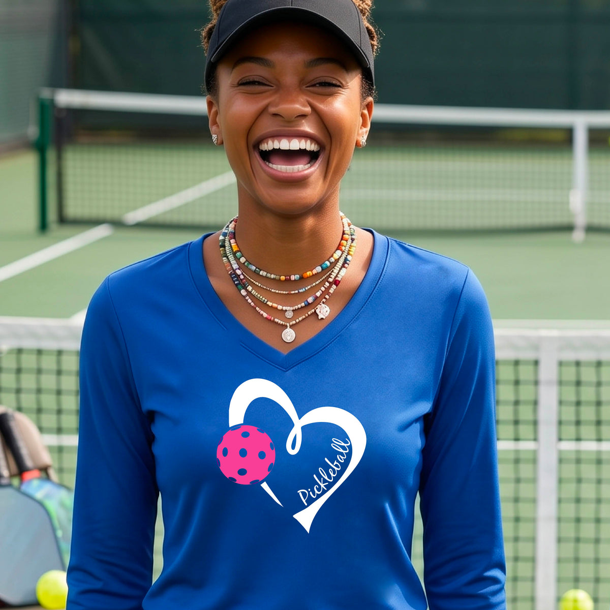 A woman smiling on a pickleball court wearing a bright blue long-sleeve V-neck athletic shirt with a white heart outline, pink pickleball graphic, and the word 'Pickleball' printed on the front. 