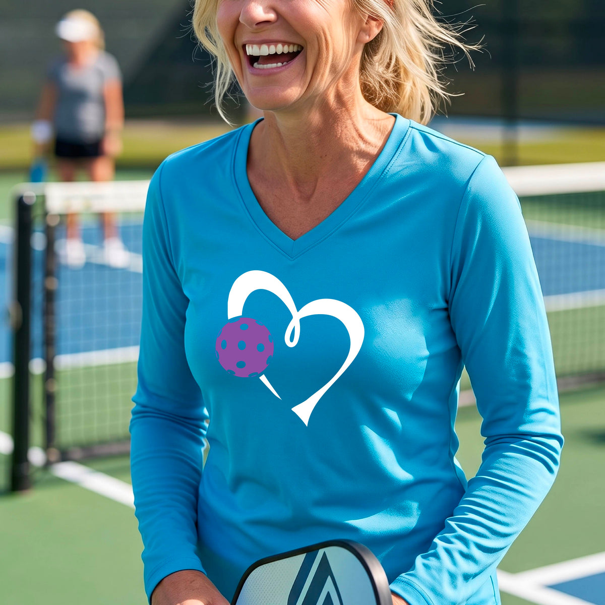 A smiling older woman on a pickleball court wearing a bright blue long-sleeve V-neck shirt with a white heart outline and a purple pickleball graphic. 