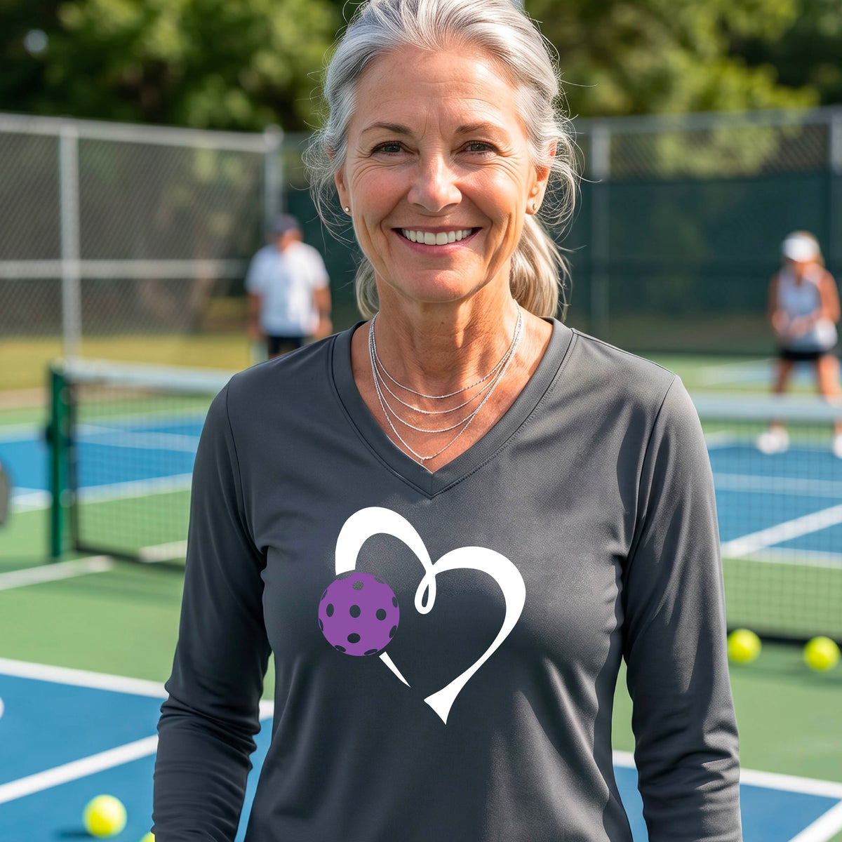 A smiling older woman on a pickleball court wearing a dark gray long-sleeve V-neck shirt with a white heart outline and a purple pickleball graphic. 