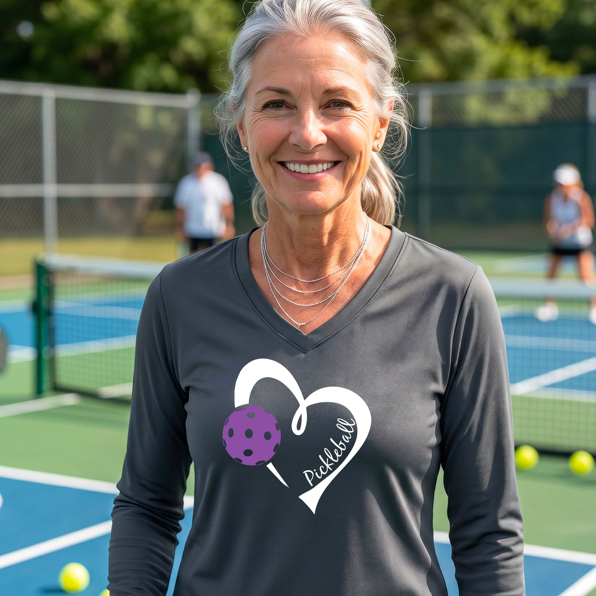 A smiling woman stands on a pickleball court wearing a dark charcoal long-sleeve V-neck shirt with a white heart outline, a purple pickleball graphic, and the word 'Pickleball' printed on the front. 