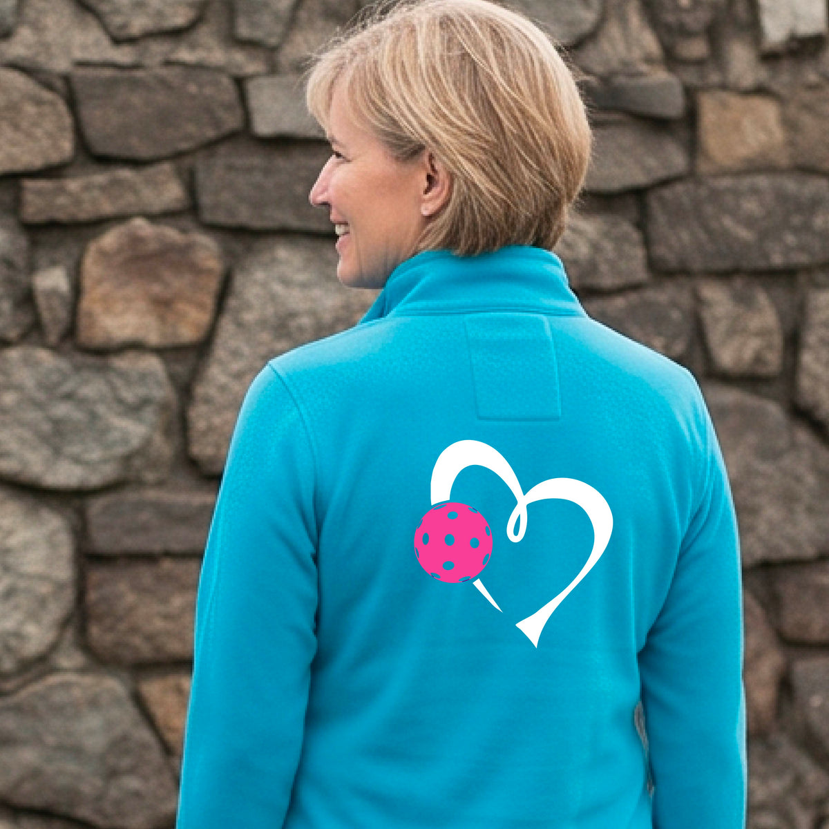 A woman on a pickleball court wearing a bright blue athletic quarter-zip shirt jacket with a white heart and pink pickleball graphic on the back.