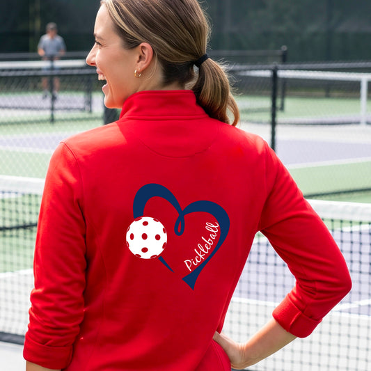A woman on a pickleball court wearing a red long-sleeve athletic quarter-zip jacket, shown from the back. The jacket features a navy blue heart outline with a white pickleball graphic and the word 'Pickleball' written inside.