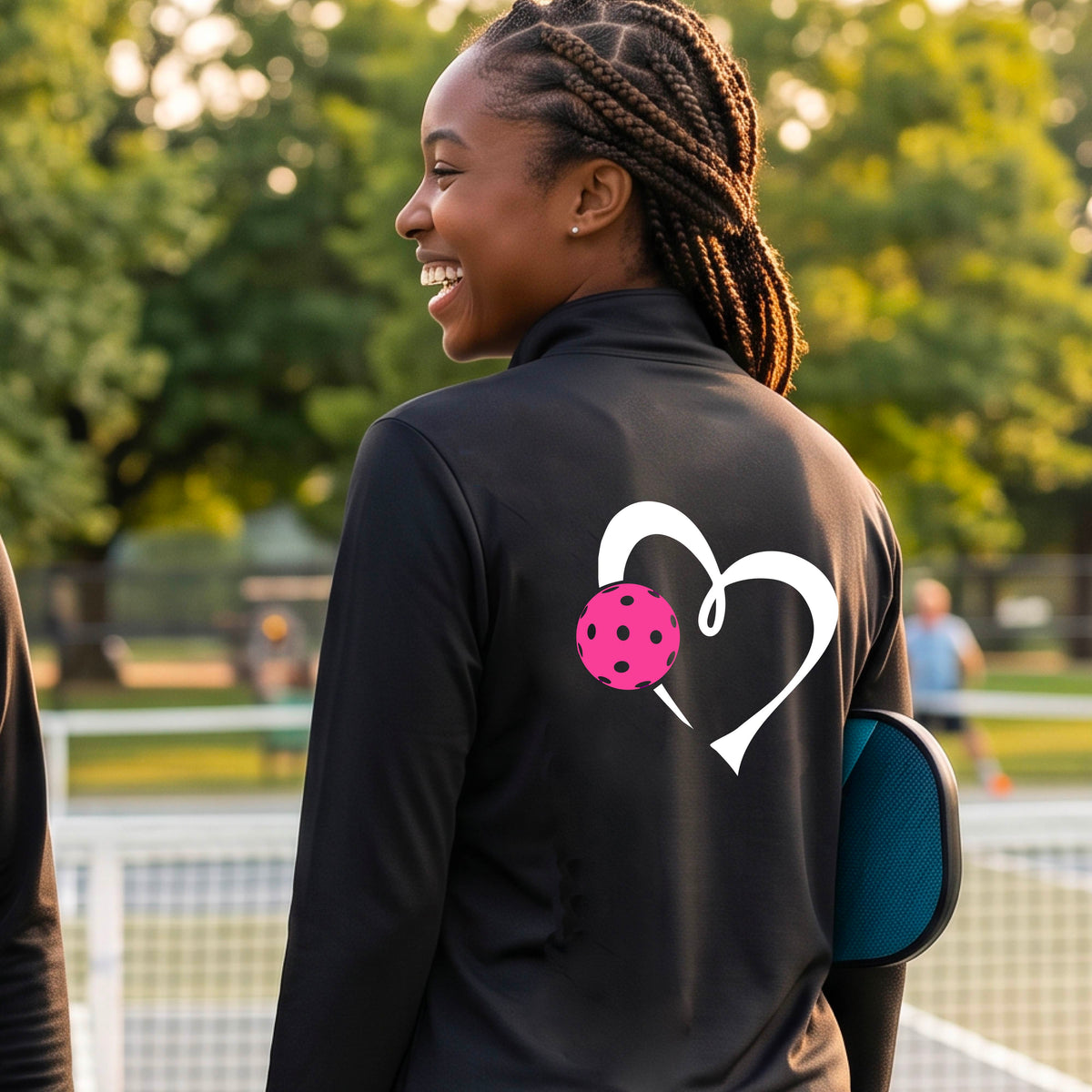 A woman on a pickleball court wearing a black athletic quarter-zip shirt jacket with a white heart and pink pickleball graphic on the back.