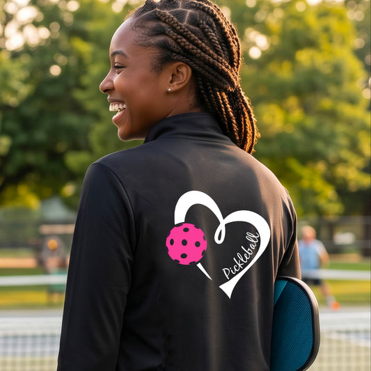 A woman on a pickleball court wearing a black athletic quarter-zip jacket with a white heart outline, a pink pickleball graphic, and the word 'Pickleball' printed on the back.