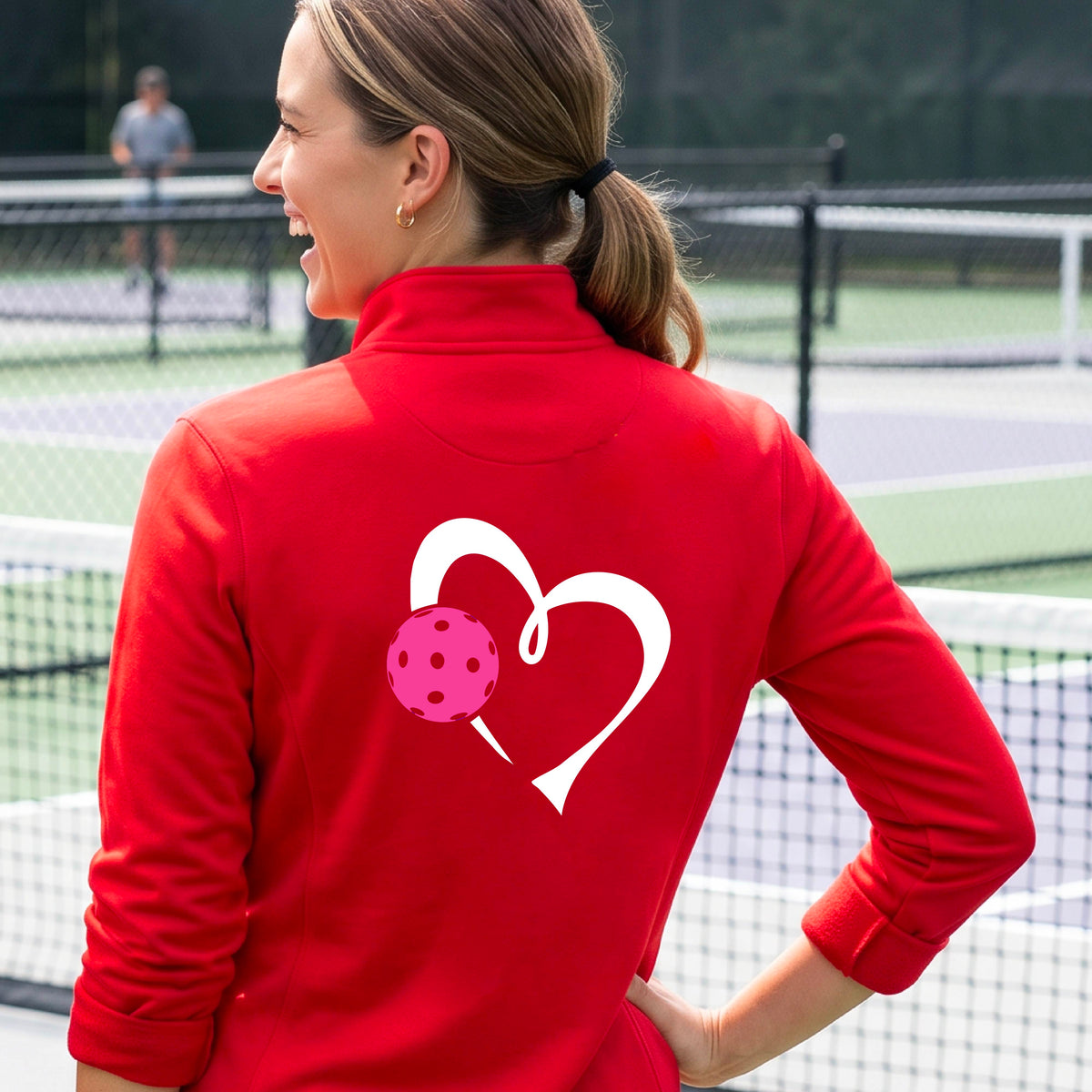 A woman on a pickleball court wearing a red athletic quarter-zip shirt jacket with a white heart and pink pickleball graphic on the back.