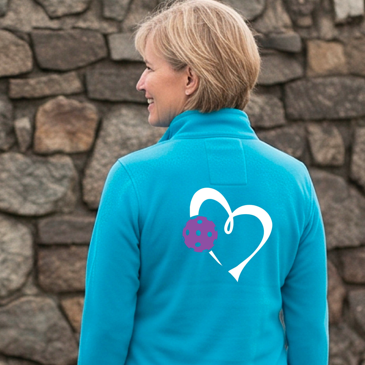 A woman wearing a bright blue quarter-zip long-sleeve athletic top, shown from the back. The top features a large white heart outline on the back with a purple pickleball graphic overlapping it.