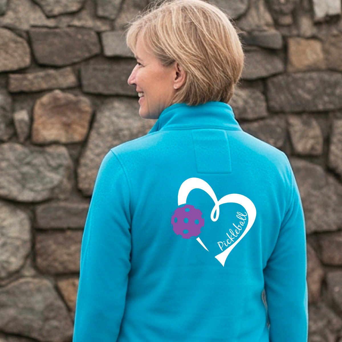 A woman on a pickleball court wearing a bright blue long-sleeve athletic quarter-zip jacket with a white heart, purple pickleball, and the word 'Pickleball' printed on the back.