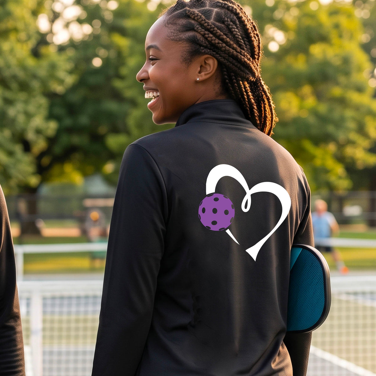A woman on a pickleball court wearing a black quarter-zip long-sleeve athletic top, shown from the back. The top features a large white heart outline on the back with a purple pickleball graphic overlapping it.