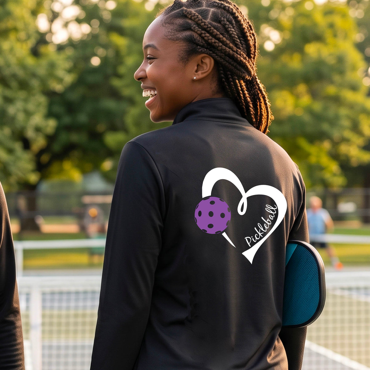 A woman on a pickleball court wearing a black long-sleeve athletic quarter-zip jacket with a white heart, purple pickleball, and the word 'Pickleball' printed on the back.