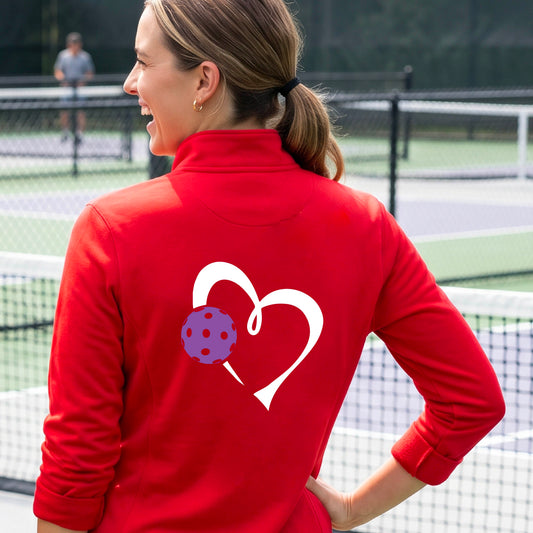 A woman on a pickleball court wearing a bright red quarter-zip long-sleeve athletic top, shown from the back. The top features a large white heart outline on the back with a purple pickleball graphic overlapping it.