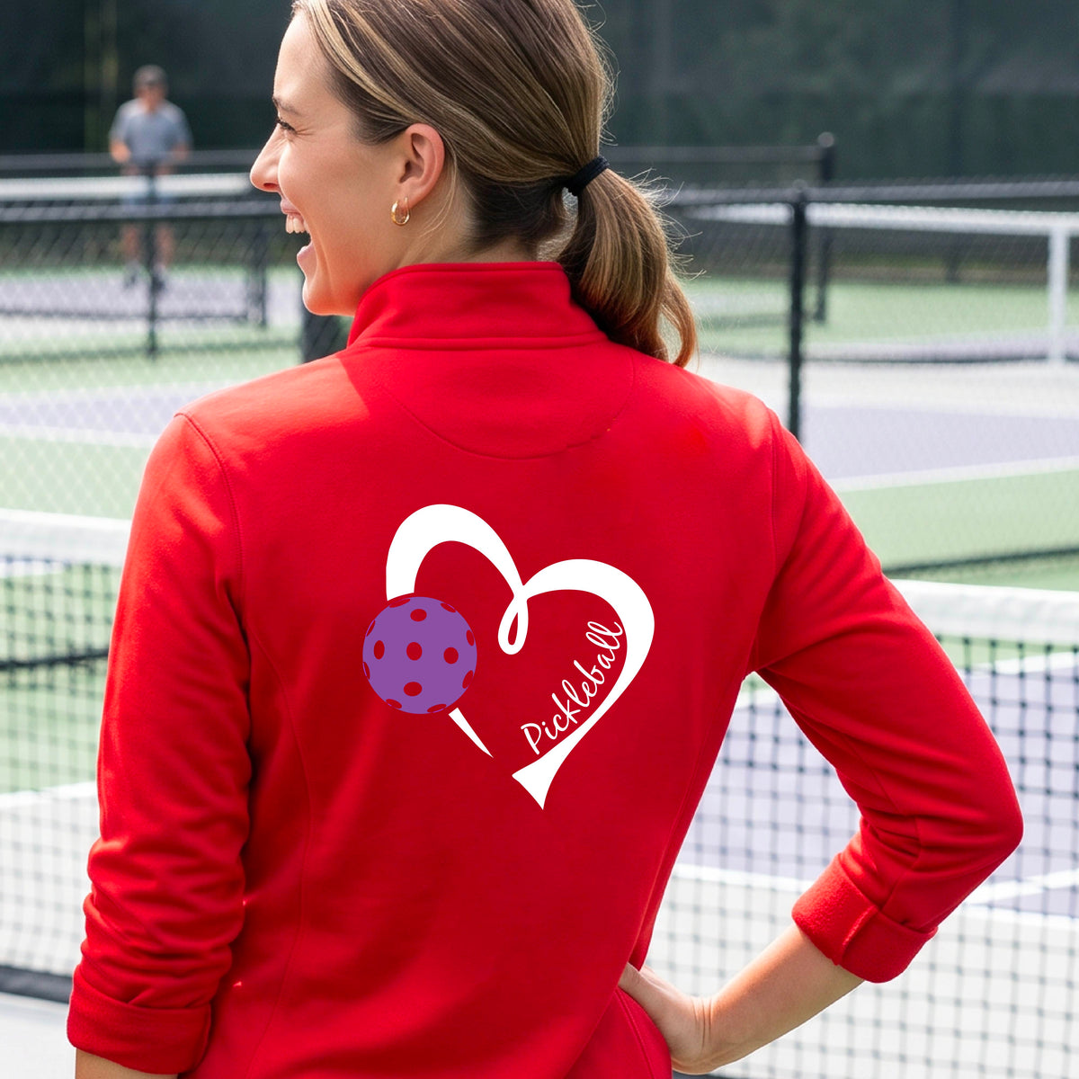 A woman on a pickleball court wearing a red long-sleeve athletic quarter-zip jacket with a white heart, purple pickleball, and the word 'Pickleball' printed on the back.