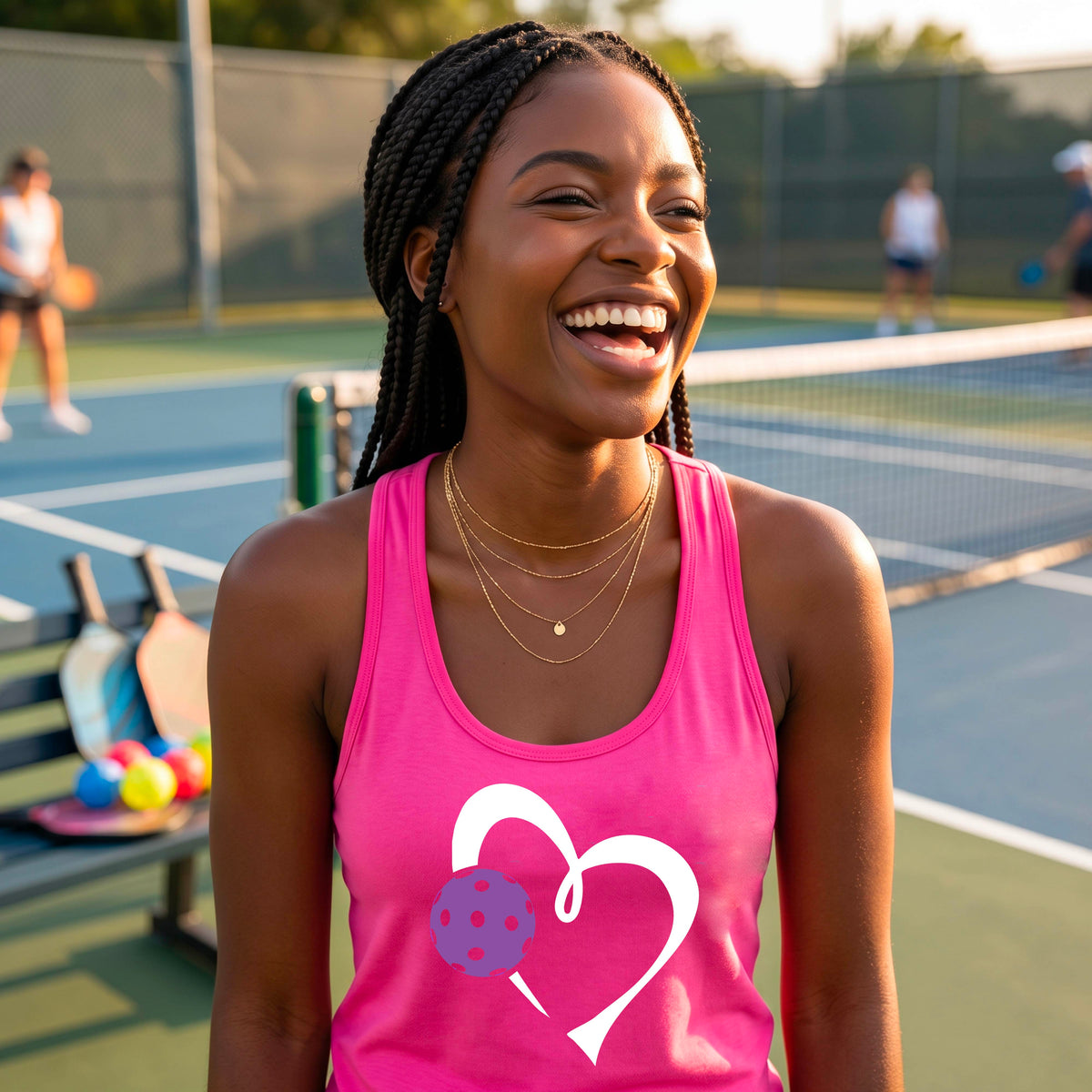 A smiling woman on a pickelball court wearing a bright pink tank top with a white heart and purple pickleball graphic, standing on a pickleball court. 