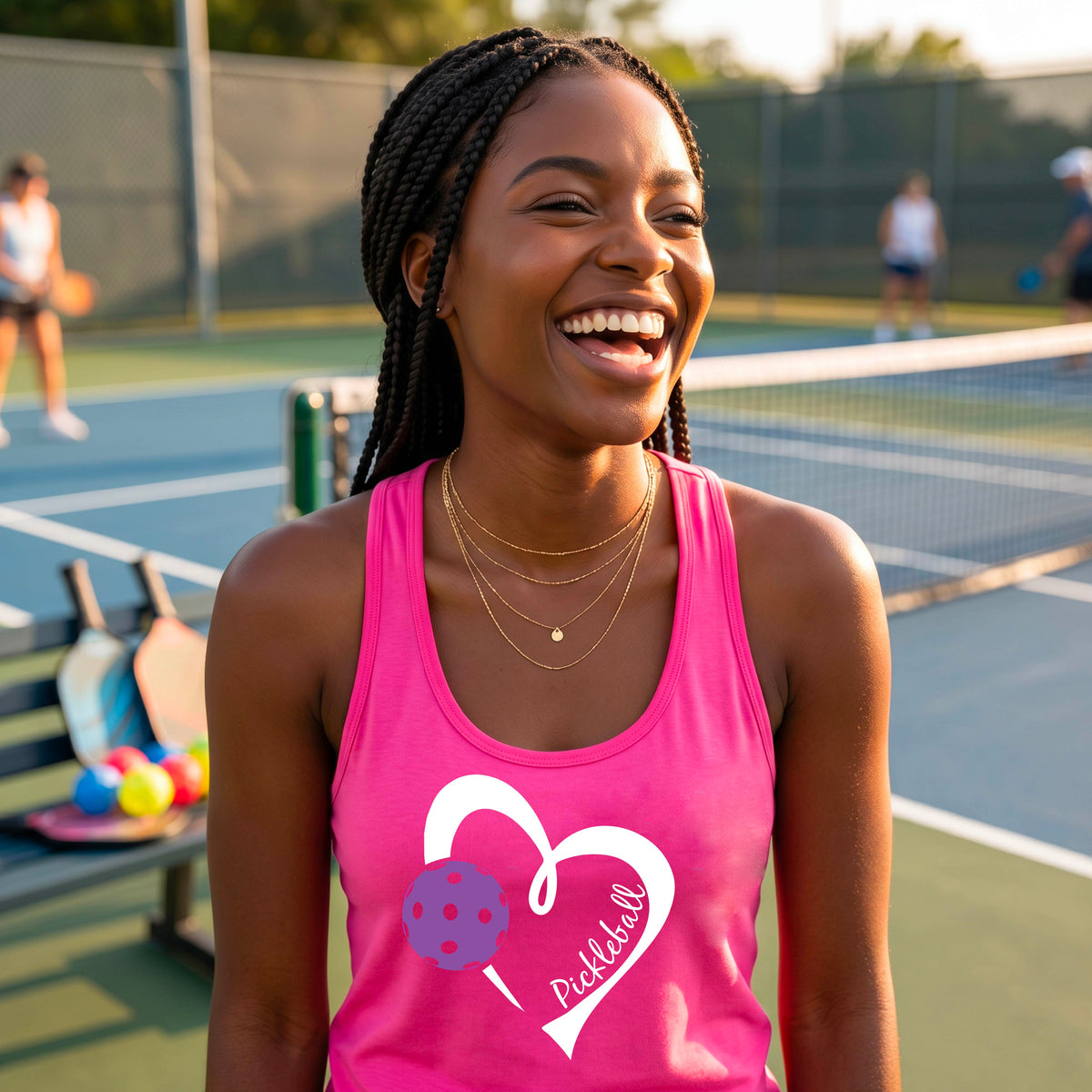 A woman smiling on a pickleball court wearing a pink tank top with a white heart outline, a purple pickleball graphic, and the word 'Pickleball' printed on the front.