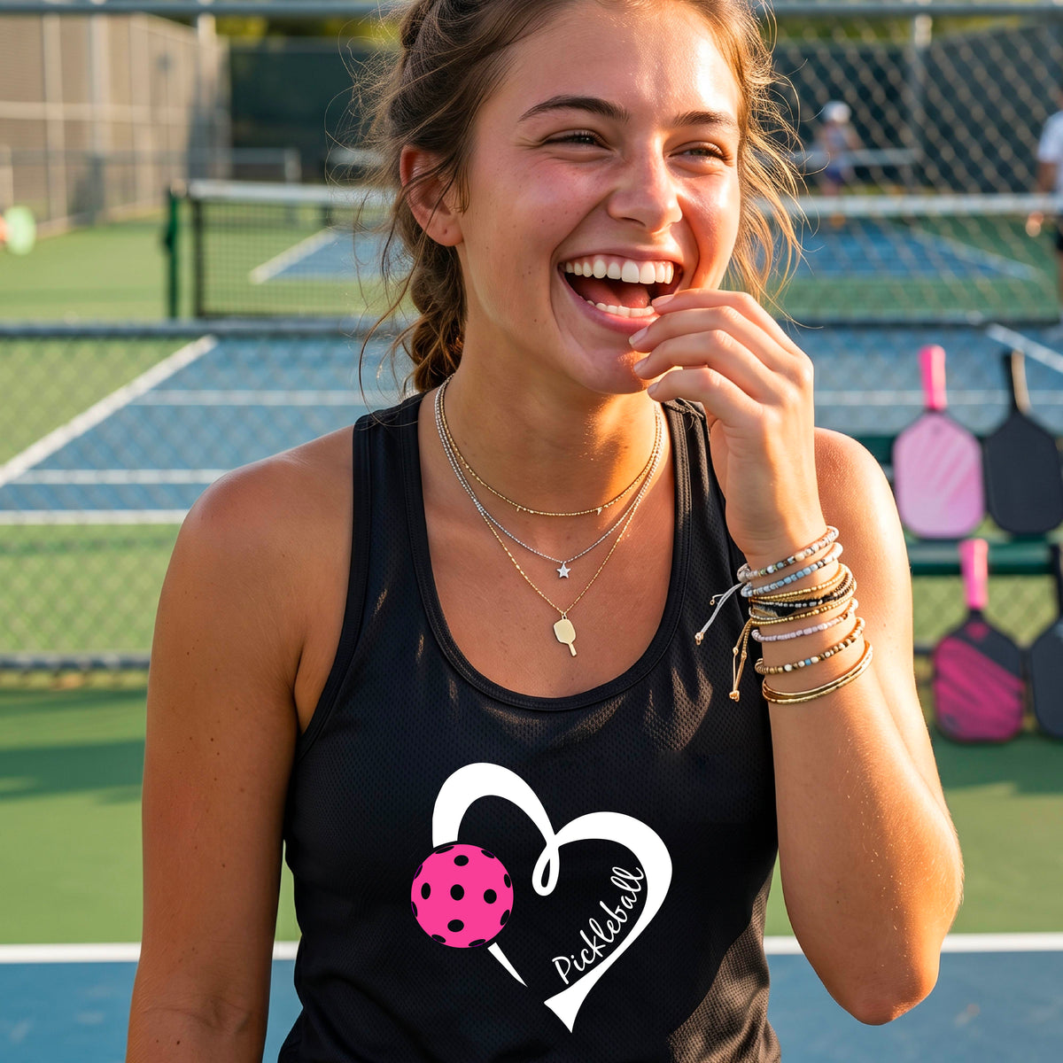 A smiling woman on a pickleball court wearing a black athletic tank top with a white heart outline, a pink pickleball graphic, and the name 'Pickball' in script. 