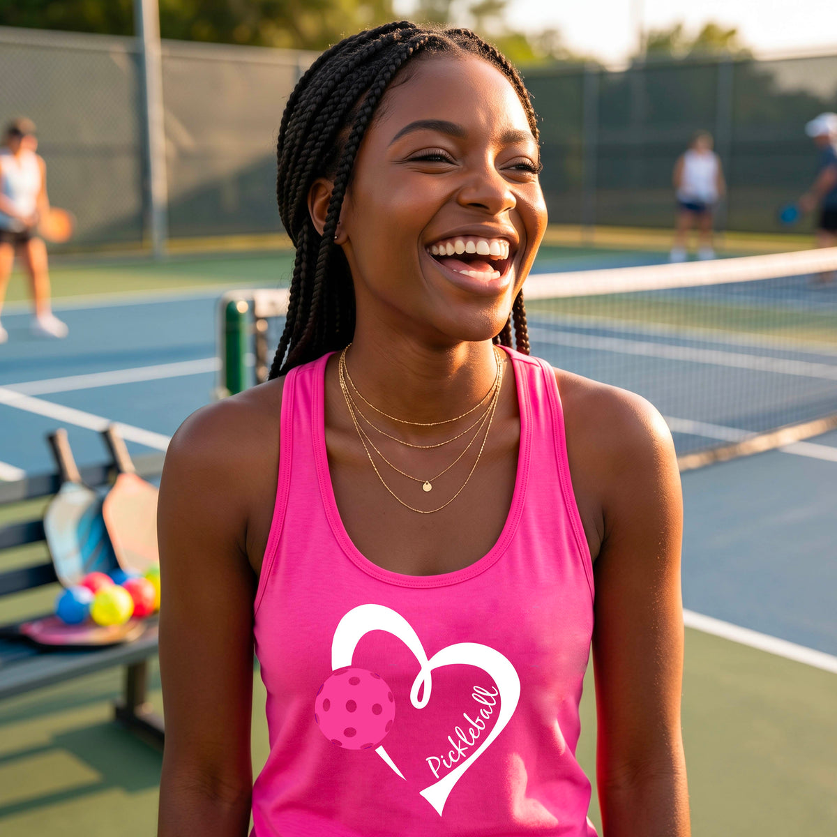A smiling woman on a pickleball court wearing a pink athletic tank top with a white heart outline, a pink pickleball graphic, and the name 'Pickball' in script. 