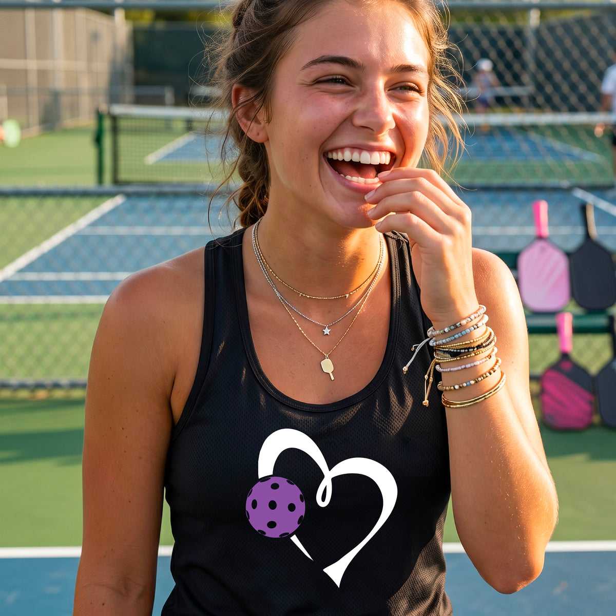 A smiling woman on a pickelball court wearing a black tank top with a white heart and purple pickleball graphic, standing on a pickleball court. 