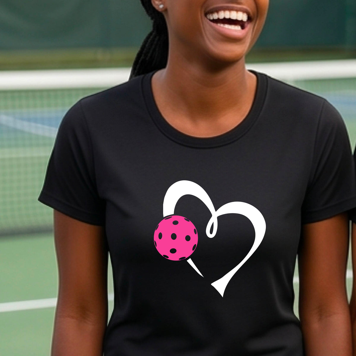 A woman wearing a black t-shirt with a white heart outline and a pink pickleball graphic on the front, set against a blurred pickleball court background.